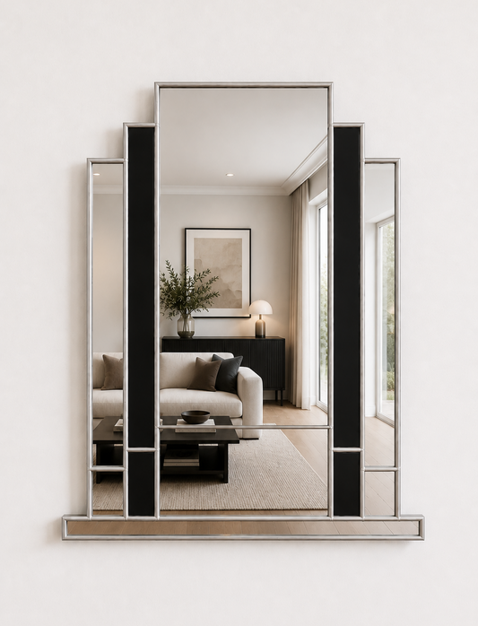 A modern geometric wall mirror with a silver-toned frame and black vertical accents, mounted on a white wall. The mirror reflects a styled living room interior.