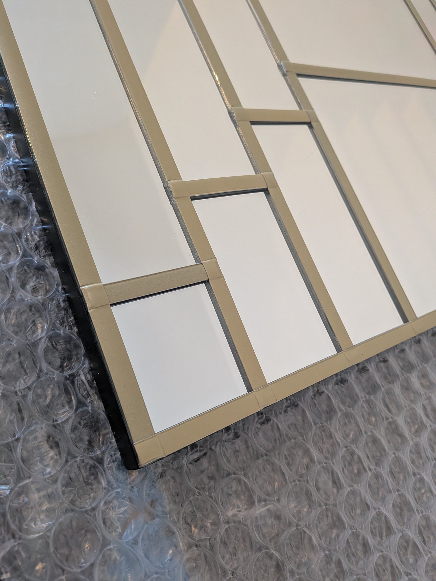 A close-up of a modern geometric wall mirror with white rectangular sections divided by a Gold frame, resting on bubble wrap packaging.