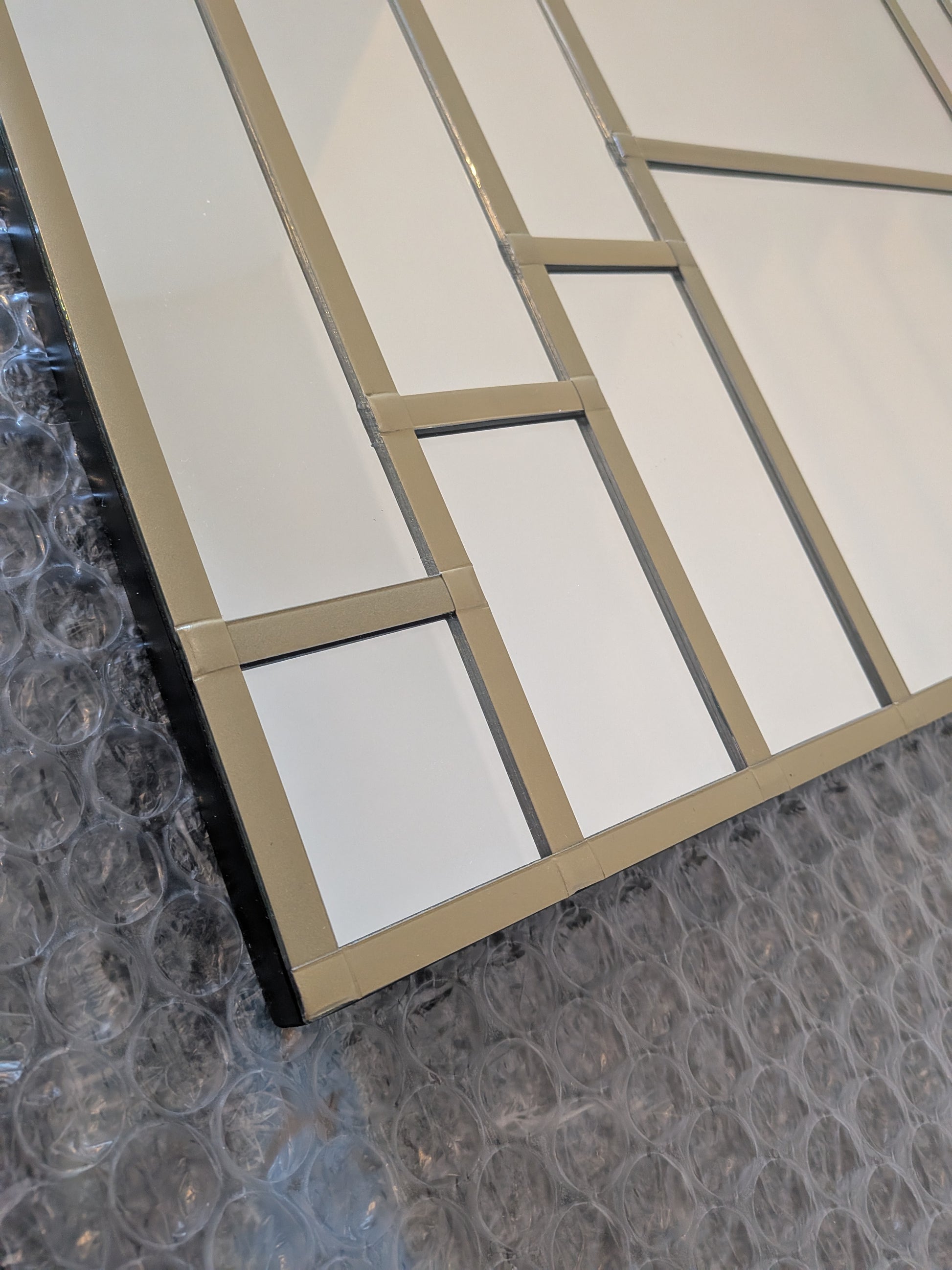 A close-up of a modern geometric wall mirror with white rectangular sections divided by a Gold frame, resting on bubble wrap packaging.